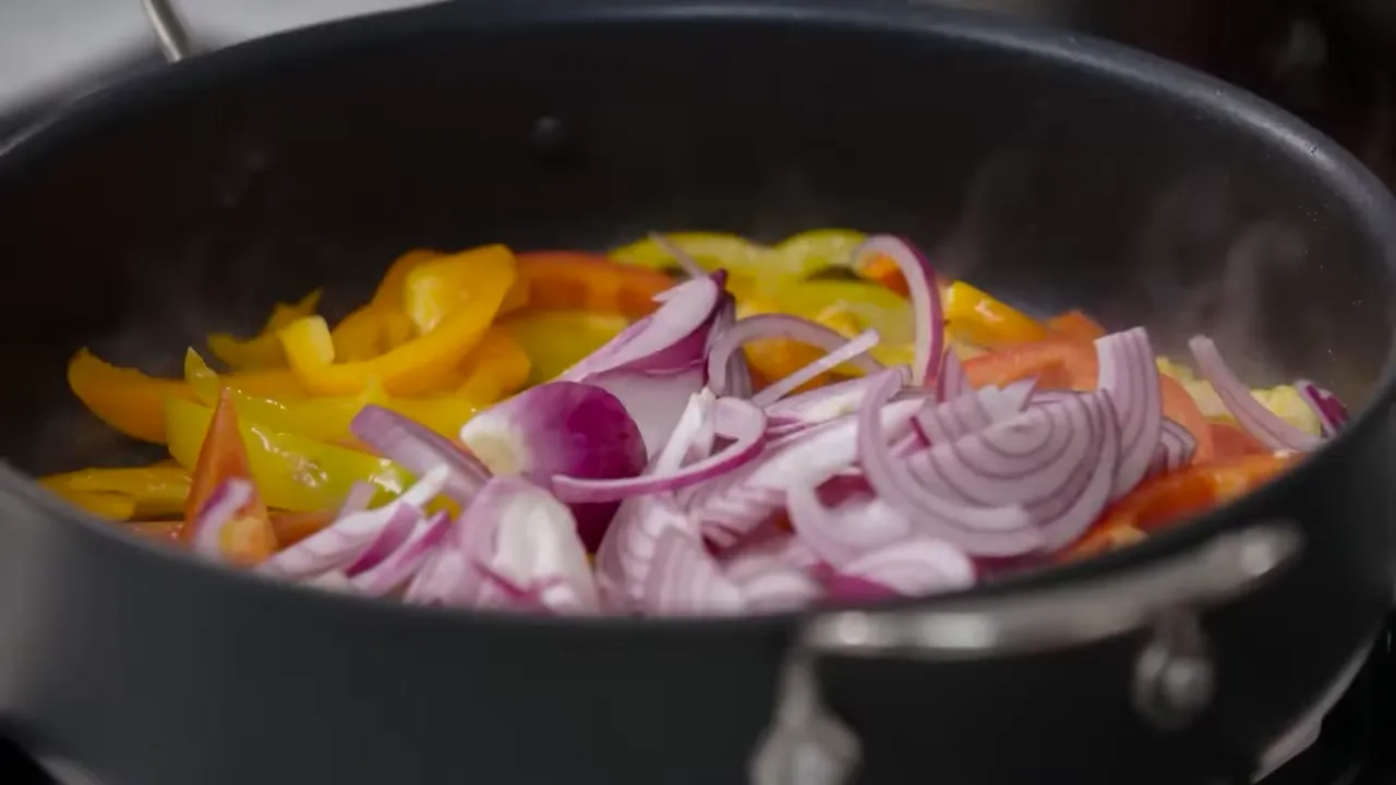 Yellow and red bell peppers with sliced red onion cooking in a large pan, visible steam and vibrant colours.