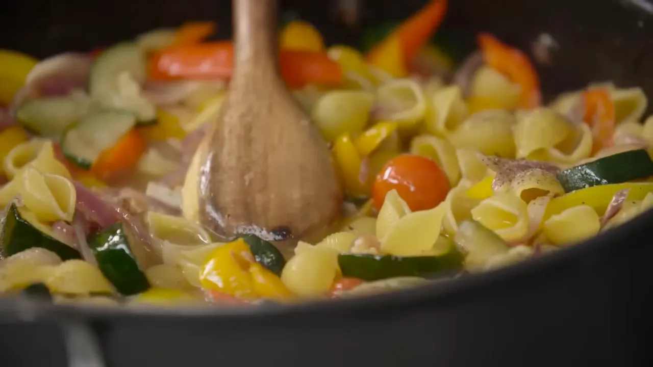Close-up of pasta shells, cherry tomatoes, courgette and peppers being stirred with a wooden spoon in a pan