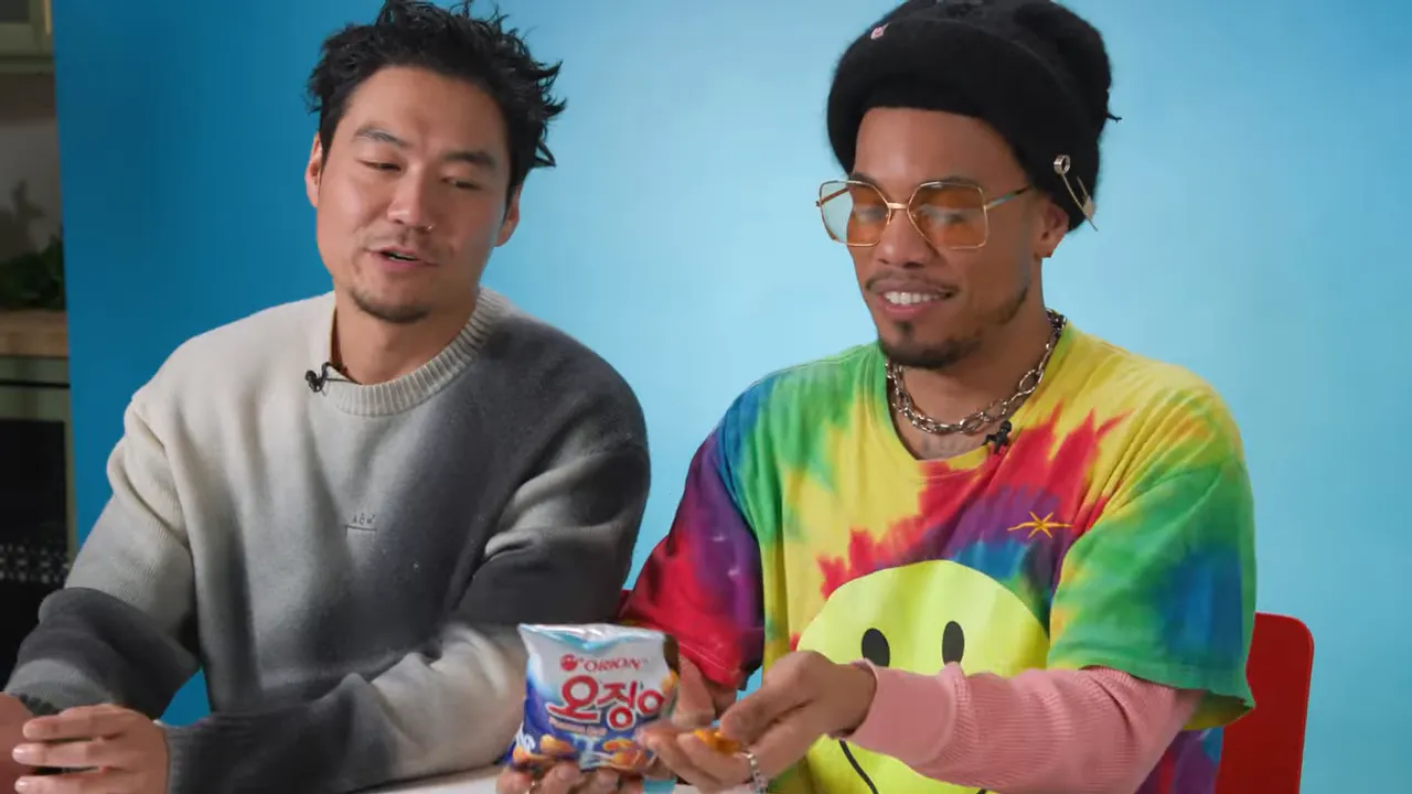 Two hosts at a table with a clear view of an Ojingeo (peanut octopus) snack bag while one host places pieces in the other's hand.