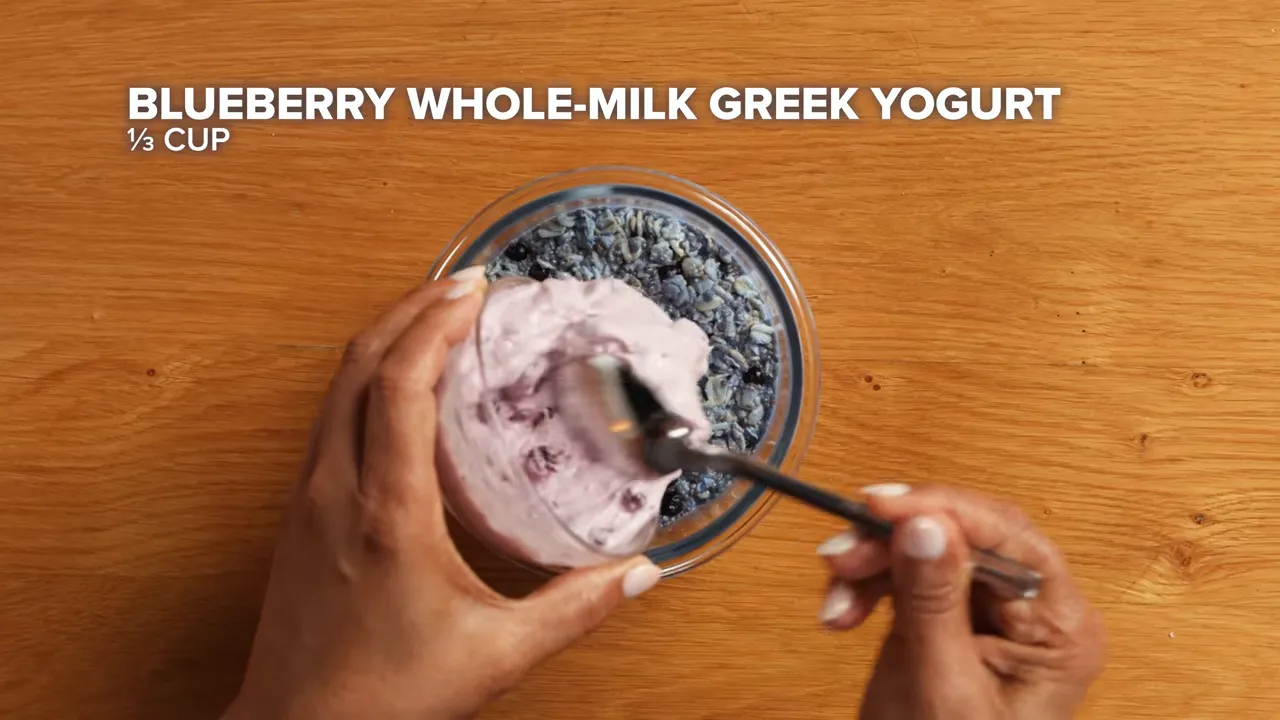 Top-down shot of a spoon adding blueberry whole-milk Greek yogurt into a jar of overnight blueberry oats.