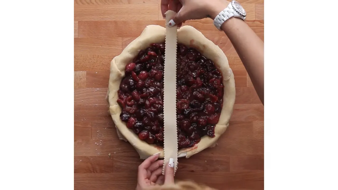 Top-down view of cherry pie filling in a crust with hands holding a pastry strip ready to place