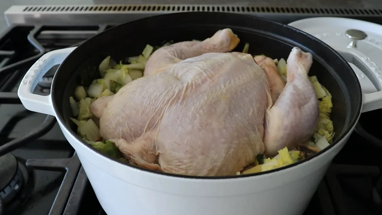 Whole chicken placed in a white Dutch oven on top of chopped onions, cabbage and other vegetables ready for simmering