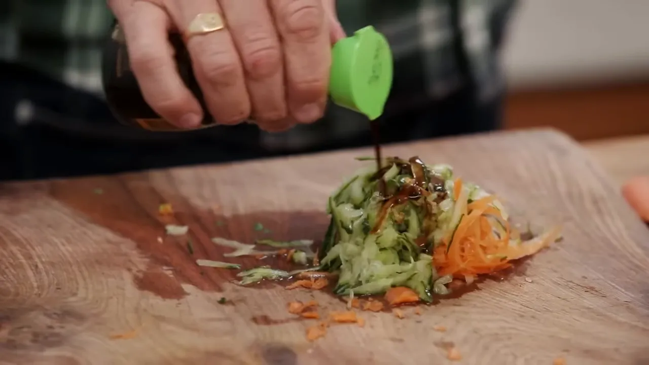 Soy sauce being poured over a pile of grated cucumber and carrot on a wooden board