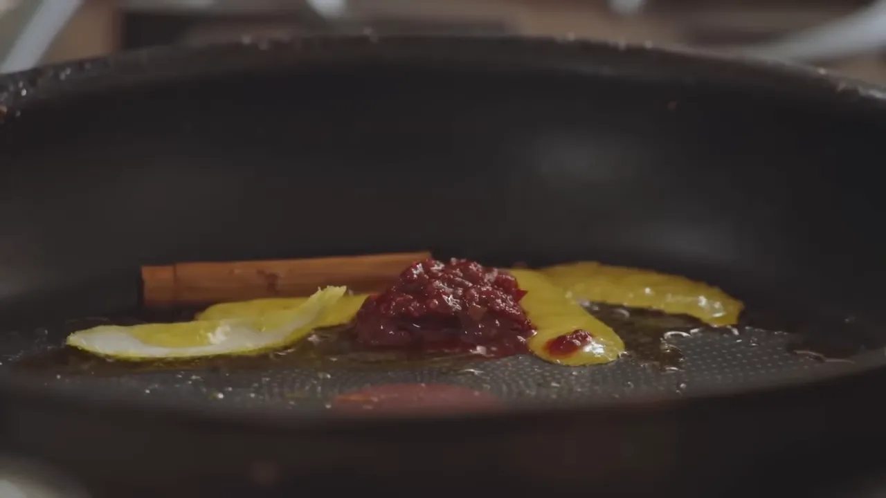 Close-up of lemon peel, a cinnamon stick and a dollop of harissa in a frying pan forming the base of a quick tomato sauce