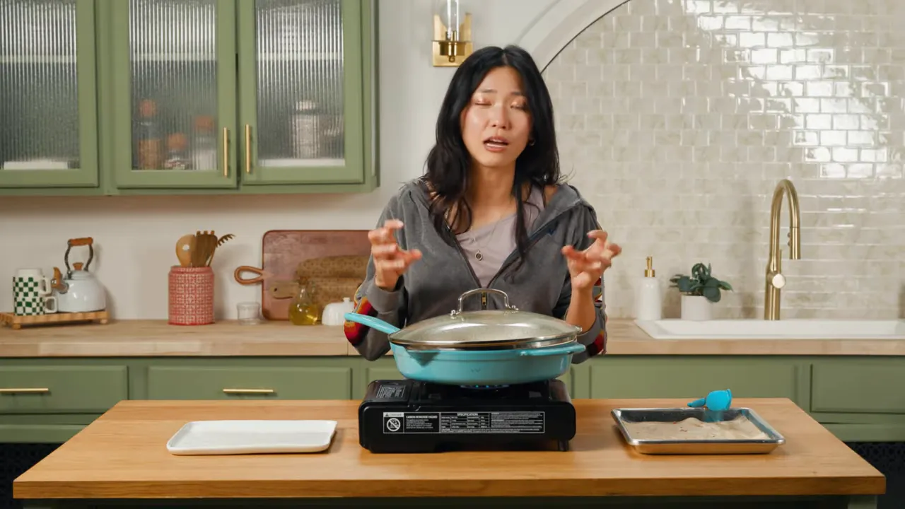 Cook standing behind countertop with covered skillet on a portable burner, trays of ingredients on either side