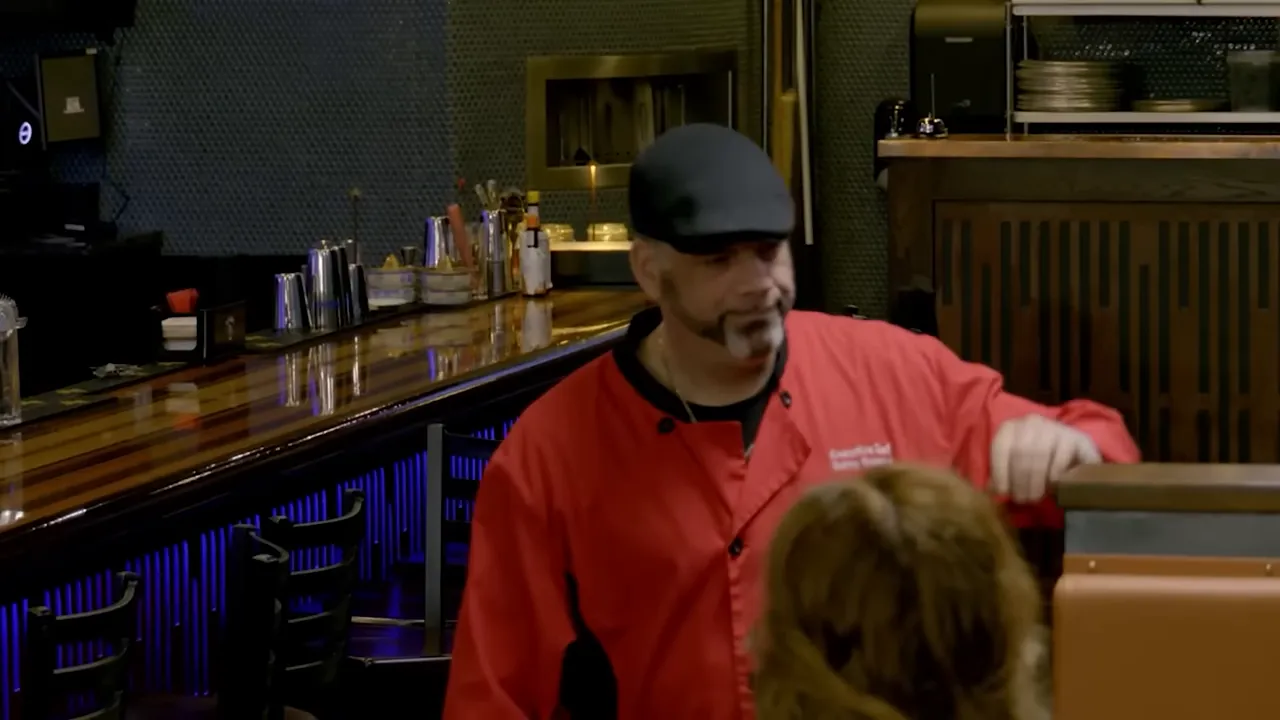 A chef or owner in a red jacket speaking with staff near the dining booths and bar.
