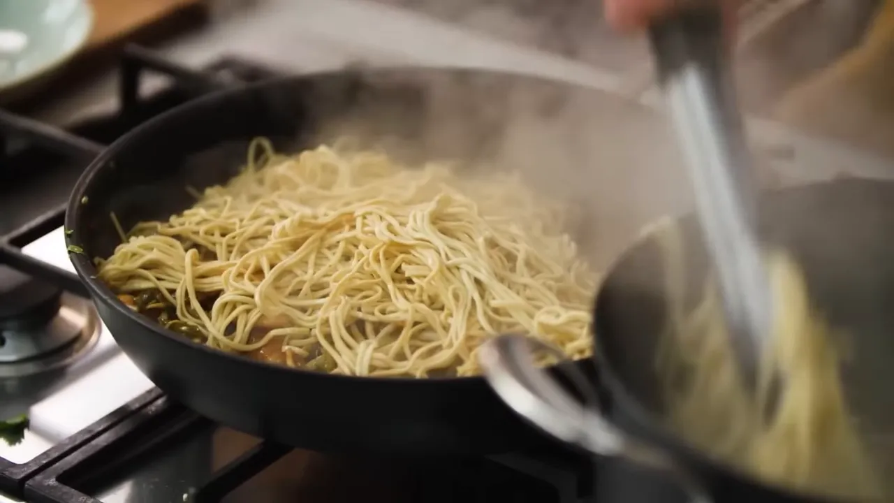 Tongs adding cooked noodles into a steaming frying pan to combine with sauce