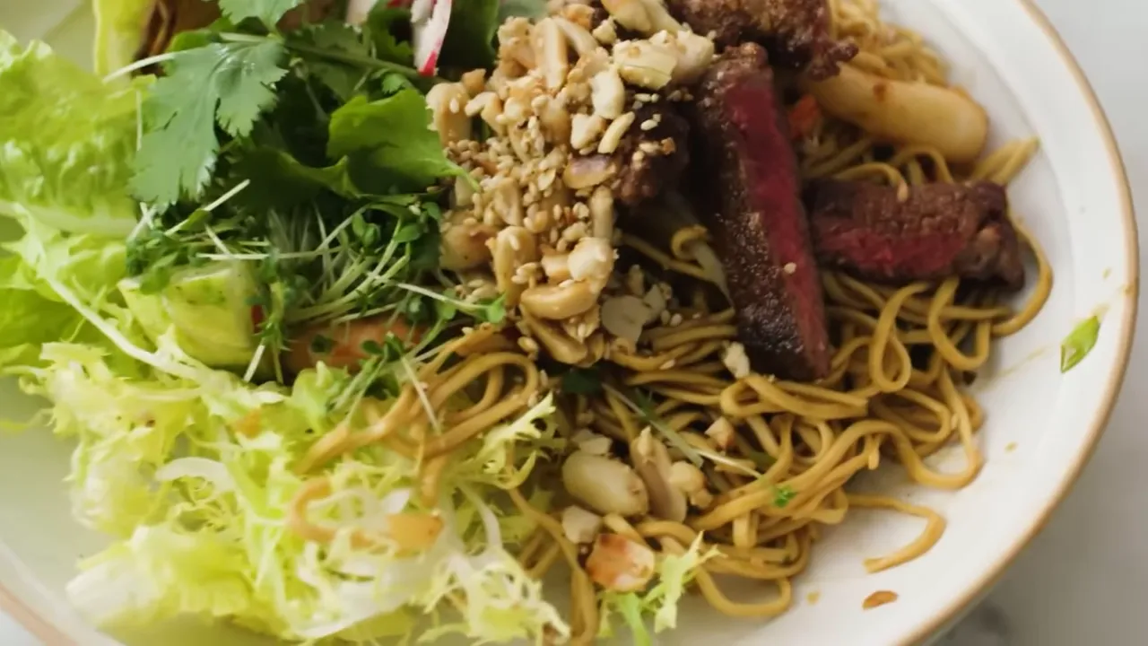 Beefsteak and hoisin prawn noodle bowl with greens, crushed nuts and noodles in a white bowl