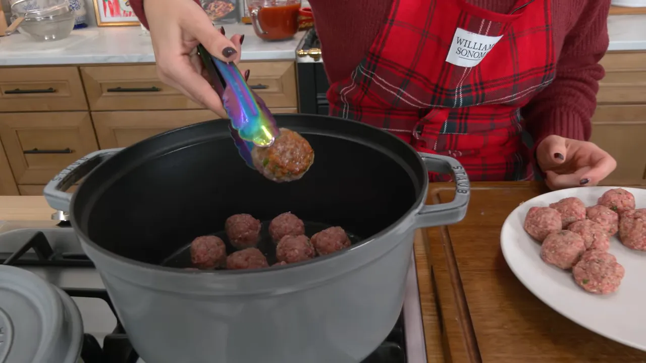 Close-up of tongs placing a meatball into a hot pot with several meatballs already inside and a plate of raw meatballs to the side.