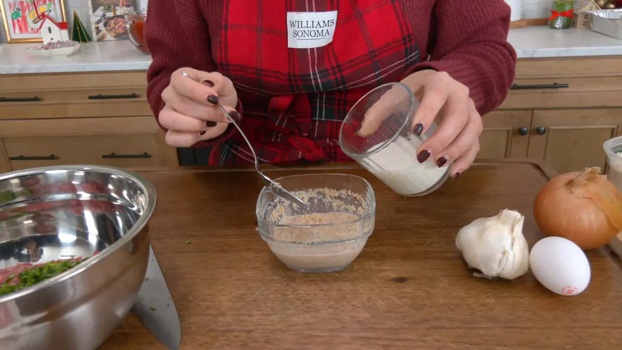 pouring milk from a small glass into a bowl of breadcrumbs on a wooden board with garlic, onion and an egg beside it
