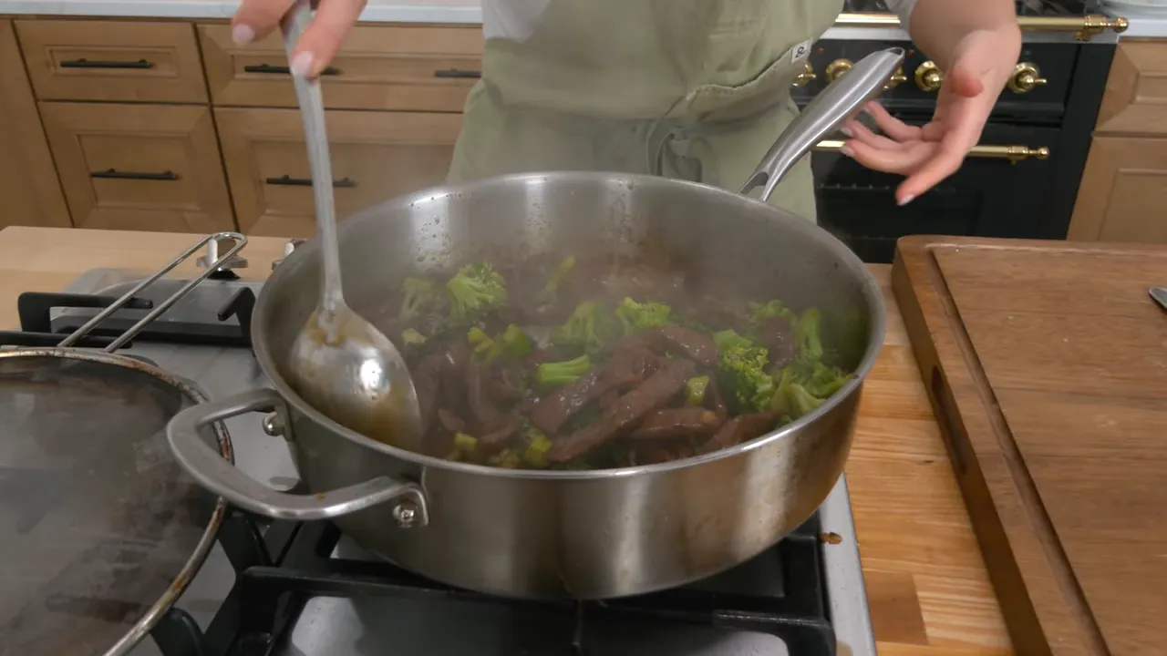 Pan of beef and broccoli in a glossy sauce being stirred with a large spoon, steam rising