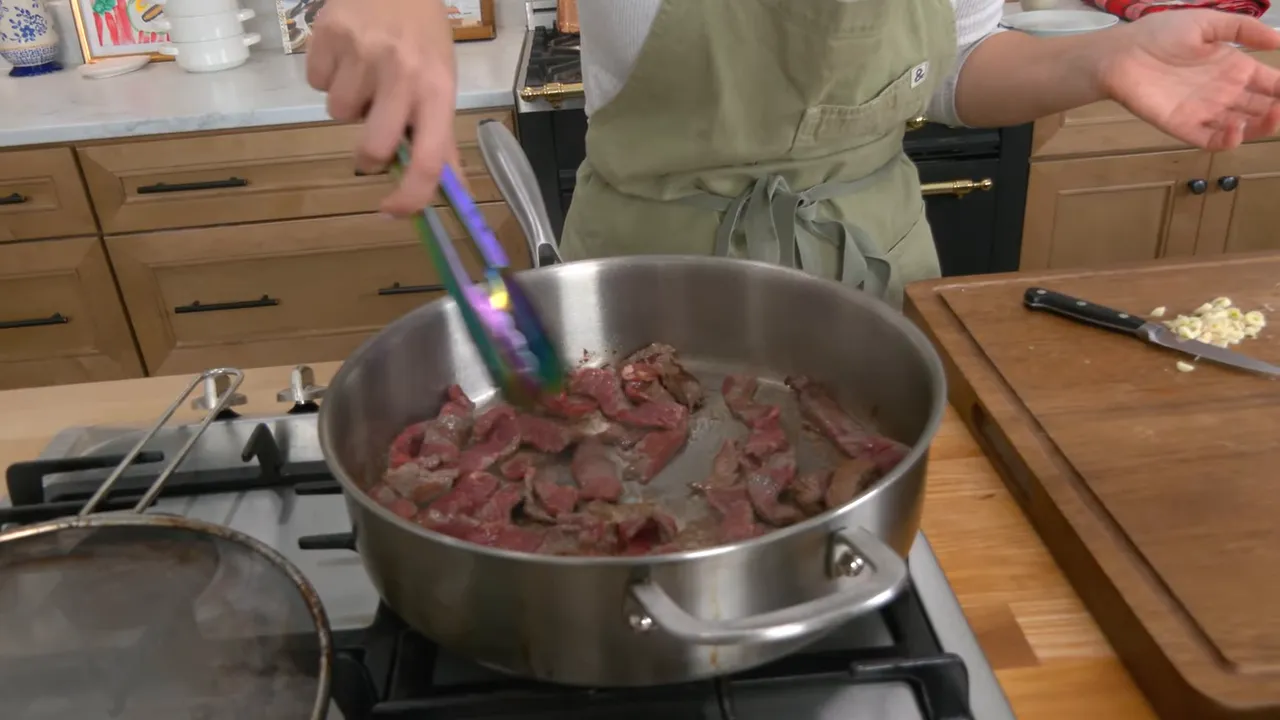 thinly sliced sirloin browning in a stainless steel pan next to a cutting board with chopped garlic