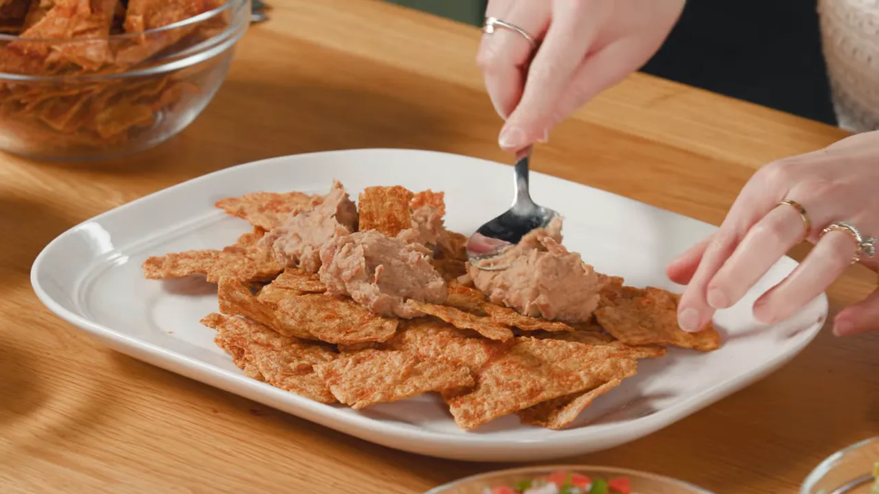 Close-up of triangular baked chicken 'chips' on a plate as refried beans are spooned on.