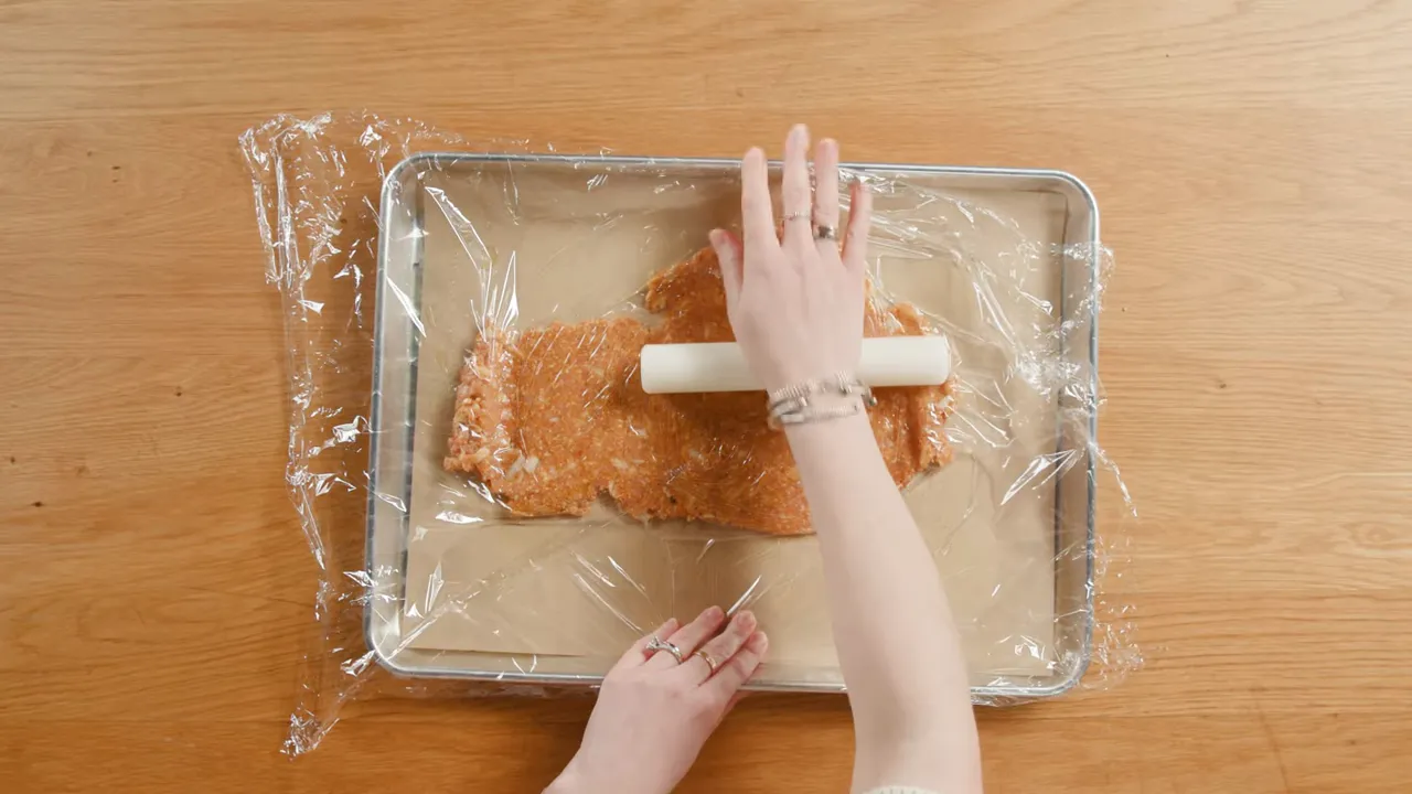 Overhead view of hands using a rolling pin to flatten ground chicken mixture under plastic wrap on a baking sheet.