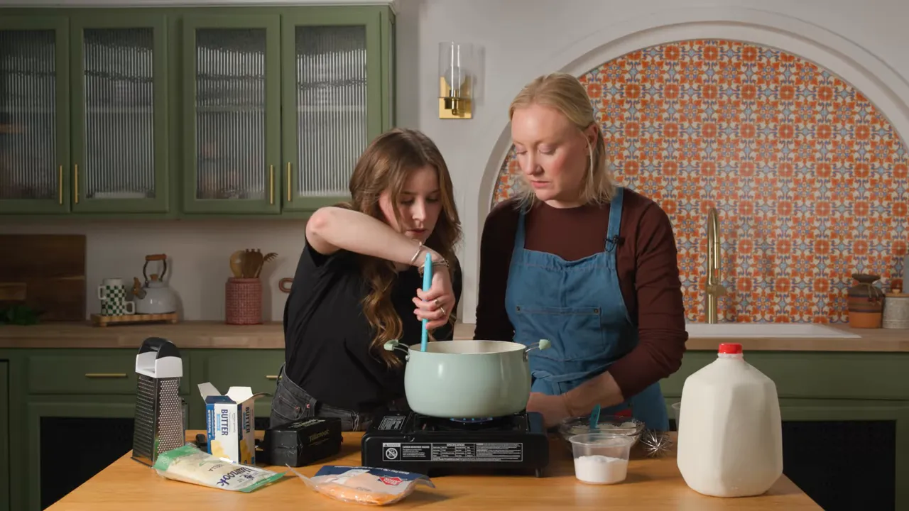 Wide frontal shot of two cooks stirring a pot on a portable burner with grated cheese, butter, milk jug and other ingredients arrayed on the countertop.