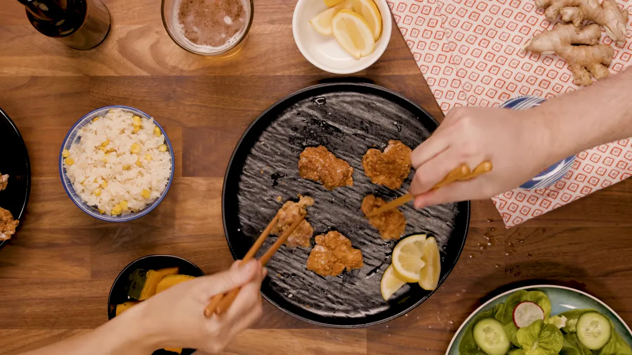 Two hands using chopsticks to take and pass karaage from a serving plate with rice and sides visible around the table