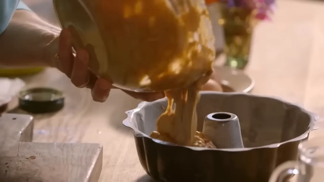 Wider shot of sticky toffee pudding batter being poured from a bowl into a bundt pan on a wooden worktop