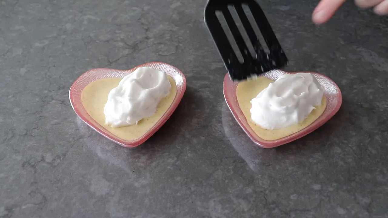 two poached meringue islands placed on vanilla crème anglaise in heart-shaped dishes being adjusted with a spatula