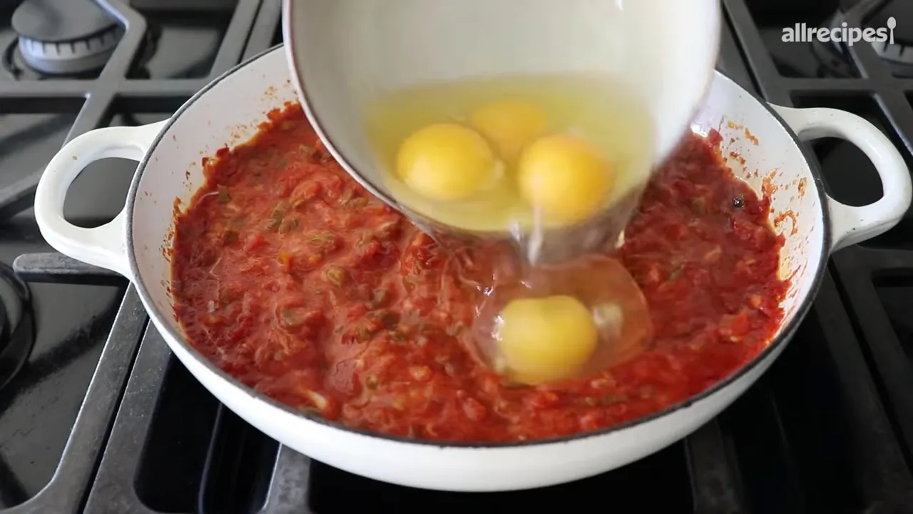 Whole eggs being poured into simmering tomato and pepper sauce in a white skillet