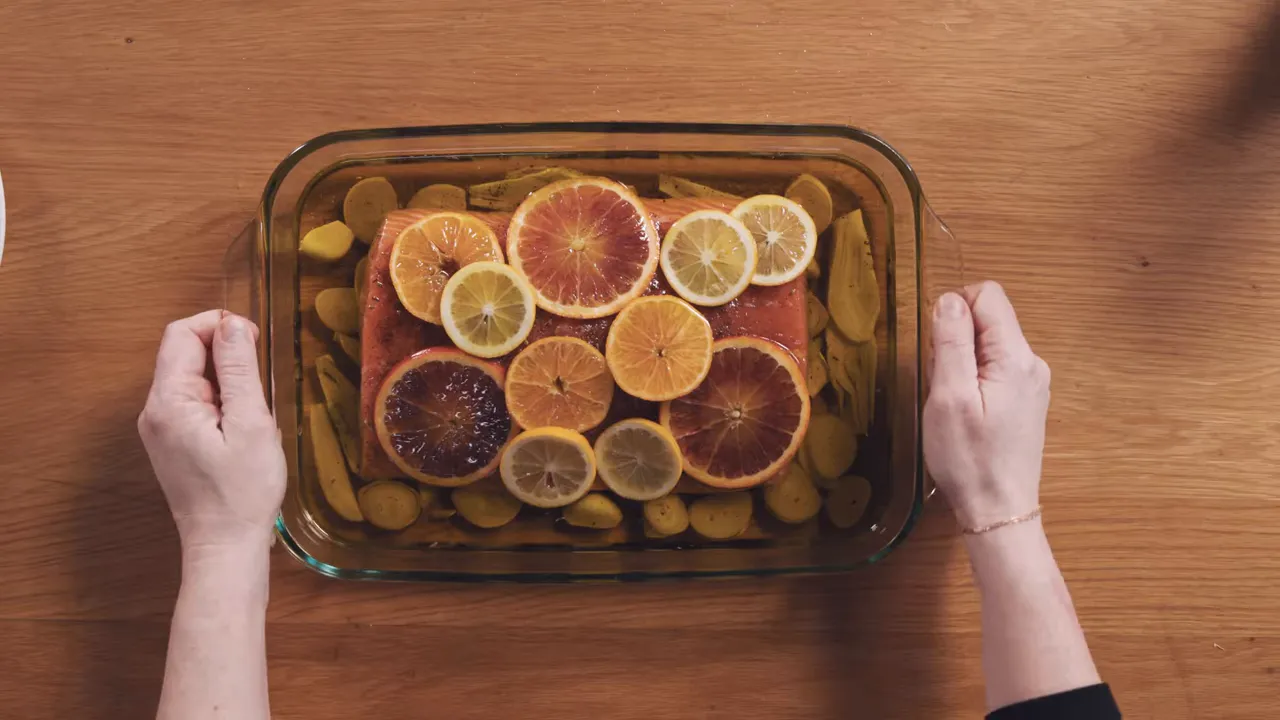 Top-down view of a salmon fillet in a glass baking dish topped with citrus slices and fennel in olive oil, with hands on either side of the dish.