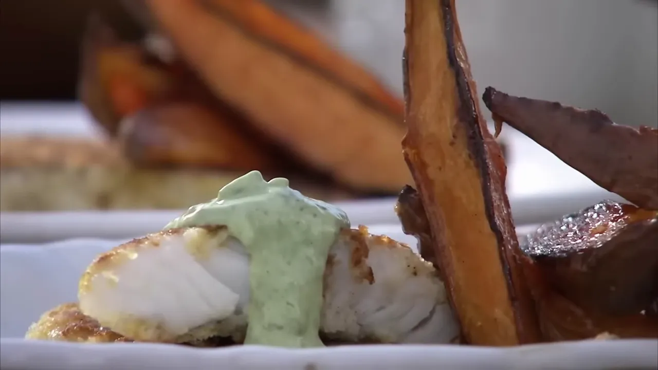 Close-up of a plated pouting fish finger with green basil mayo draped over it and roasted sweet potato wedges beside it