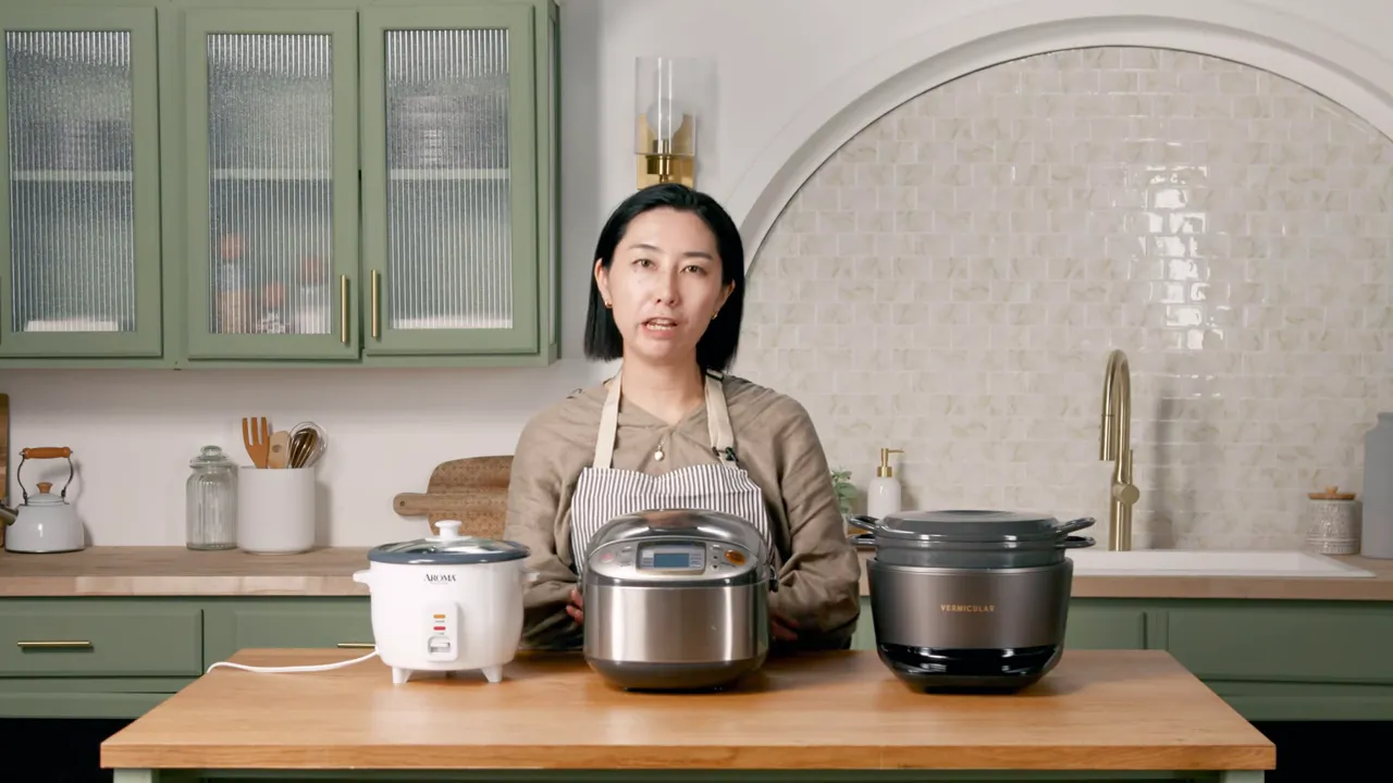 Three rice cookers lined up on a wooden table (basic white cooker, stainless multicooker, and a Vermicular unit) with the presenter behind them.