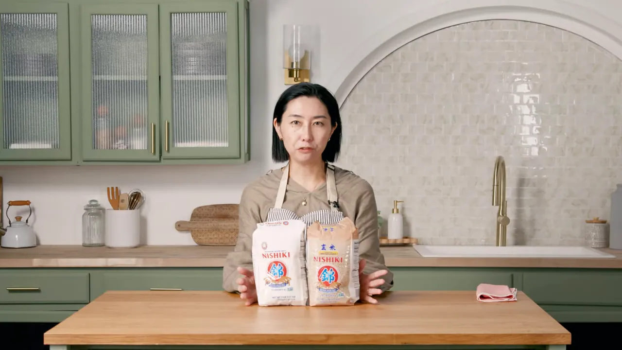 Presenter holding two bags of rice (white and brown) on a kitchen counter.