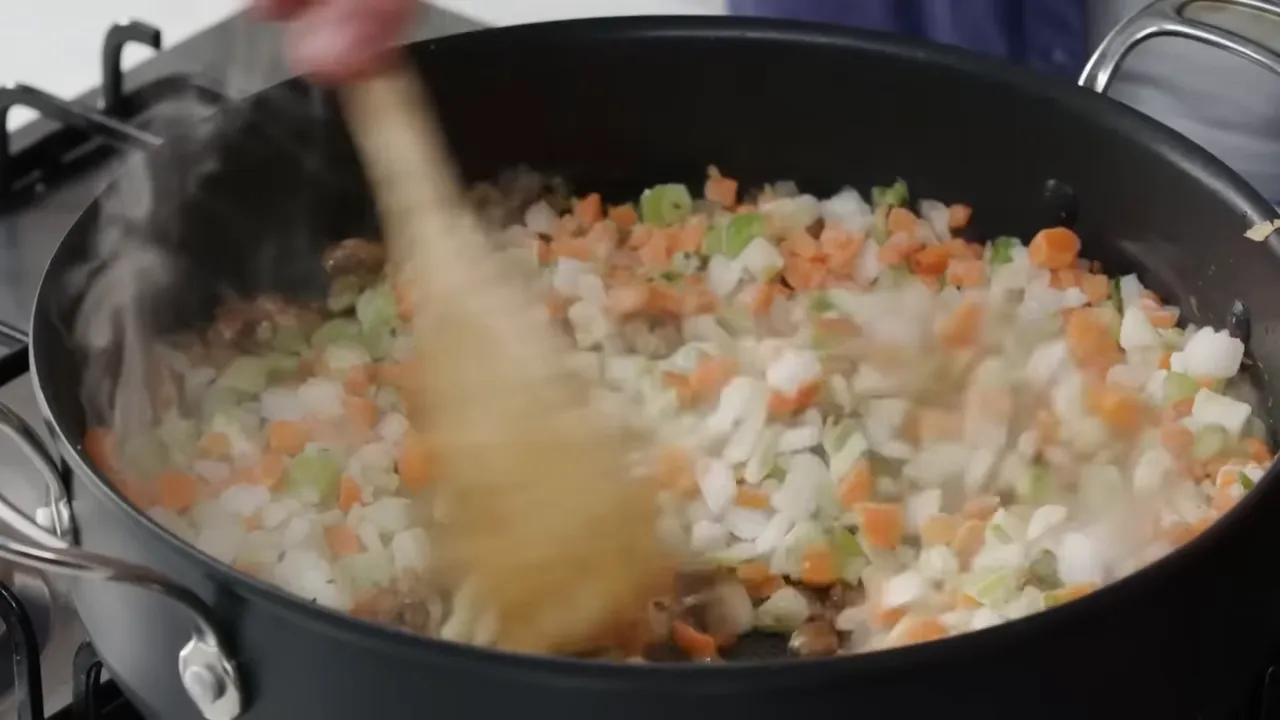 Close-up of diced carrots, onions and celery being stirred in a frying pan while steam rises.