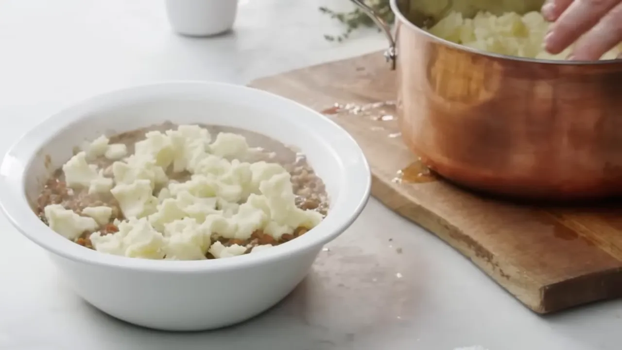 White baking dish of shepherd's pie filling topped with dollops of mashed potato beside a copper pot on a chopping board.