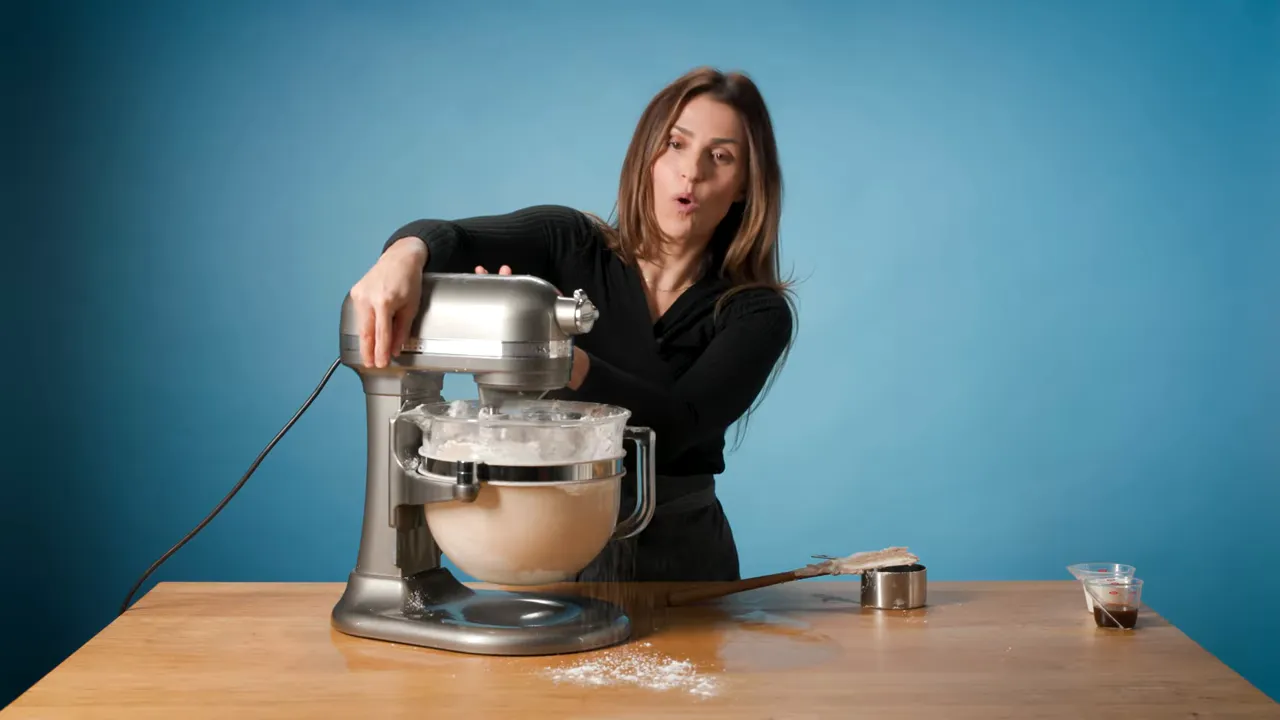 stand mixer on wooden table with spatula and measuring cup, presenter in background