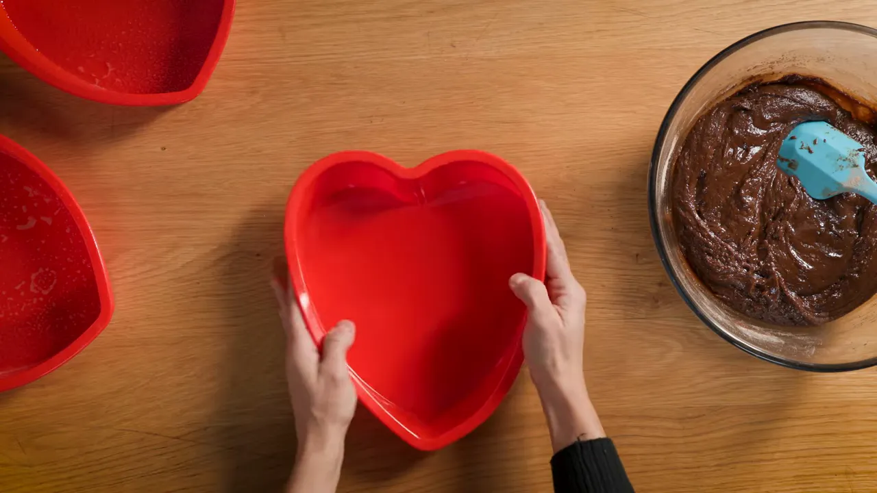 Overhead shot of a red heart-shaped silicone mold being held over a wooden table with a bowl of mocha cake batter and two additional heart molds nearby
