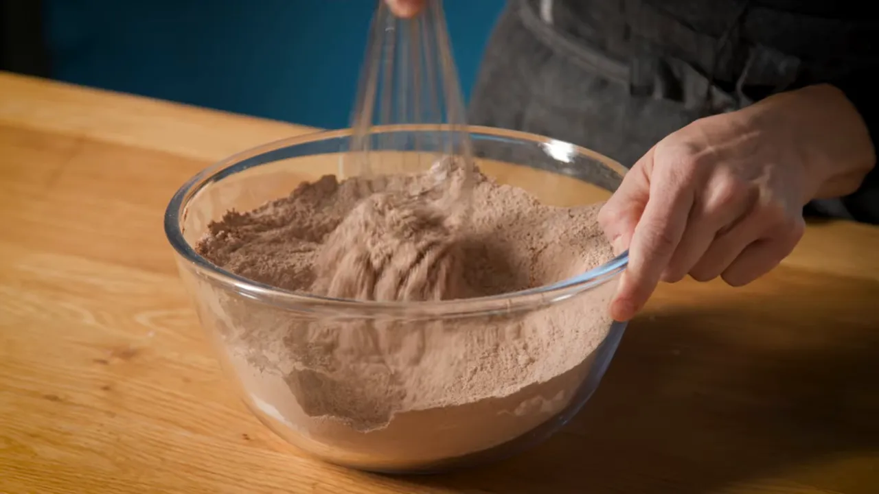 Whisk mixing dry cocoa powder, flour and espresso powder in a glass bowl on a wooden counter