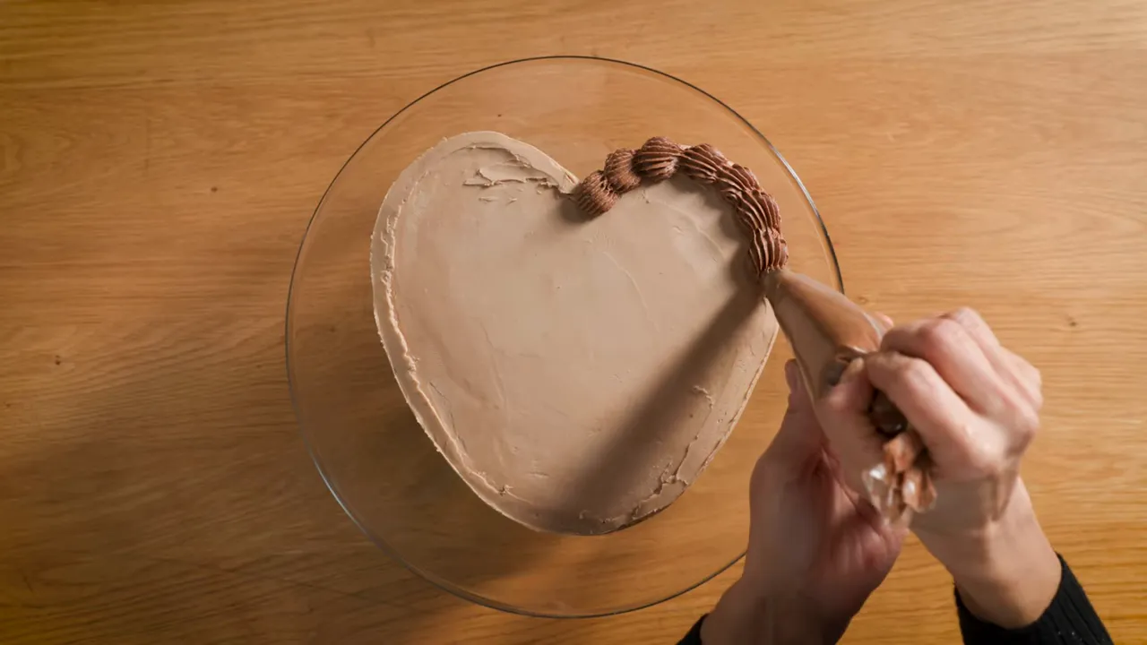 Overhead shot of a heart-shaped cake being piped with mocha buttercream along the perimeter, hands visible holding the bag