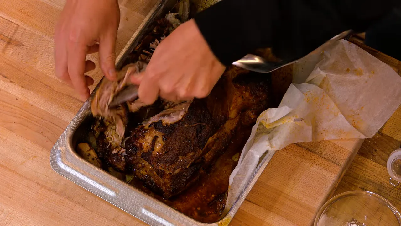 hands shredding a slow-roasted pork shoulder inside a roasting pan with parchment and juices
