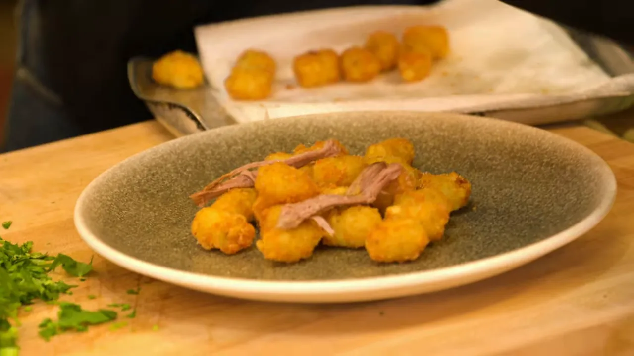 Shredded pork being placed on a bed of crispy tater tots on a plate, with herbs on the board