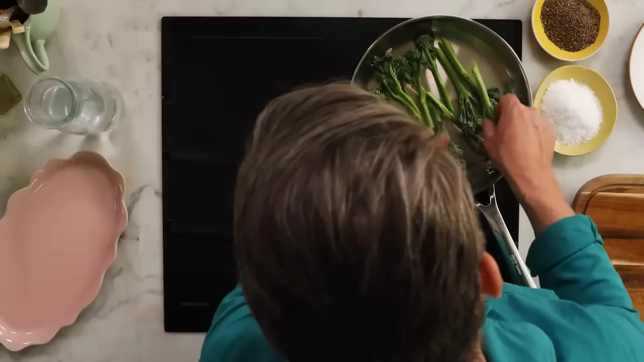 Top-down shot of a cook placing broccolini in a stainless pan on an induction hob with seasoning bowls nearby.