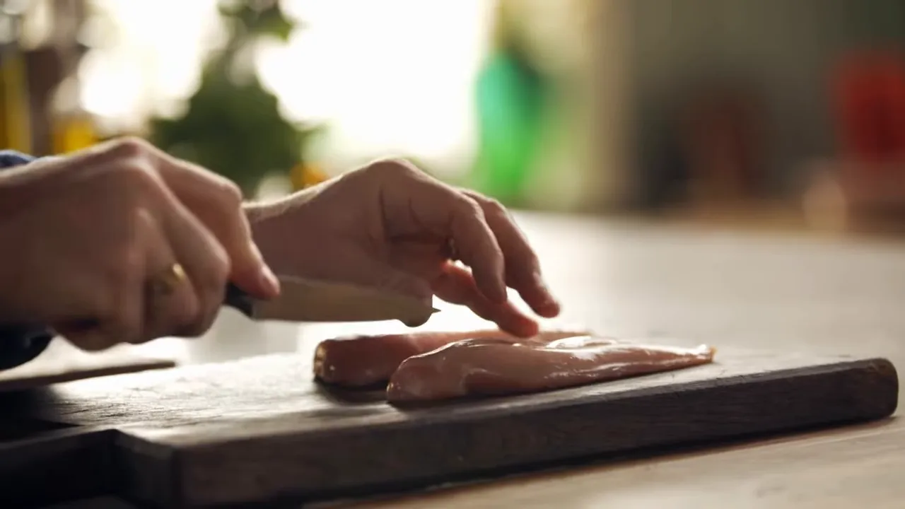 Close-up of hands scoring a chicken breast with a knife on a wooden cutting board