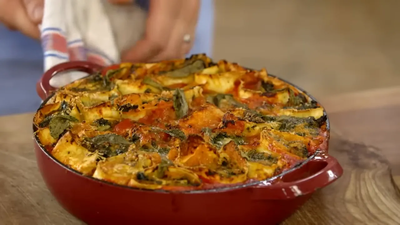Finished squash and spinach rotolo in a red baking dish, golden and bubbling with crisped sage
