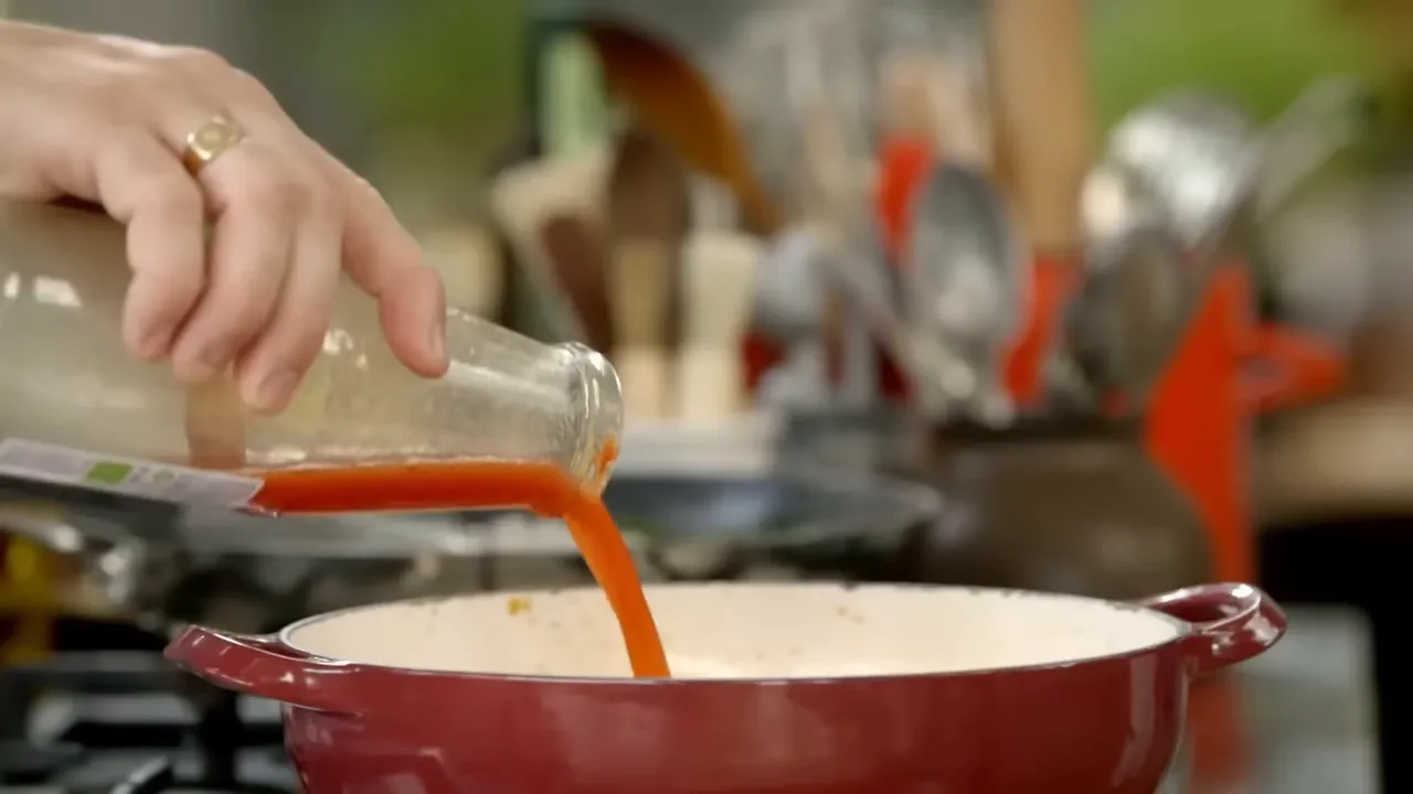 Hand pouring passata from a bottle into a red pan