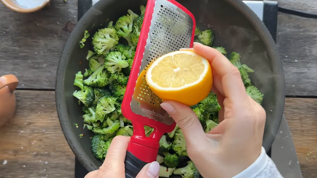 Grating lemon zest over broccoli florets in a skillet using a handheld grater