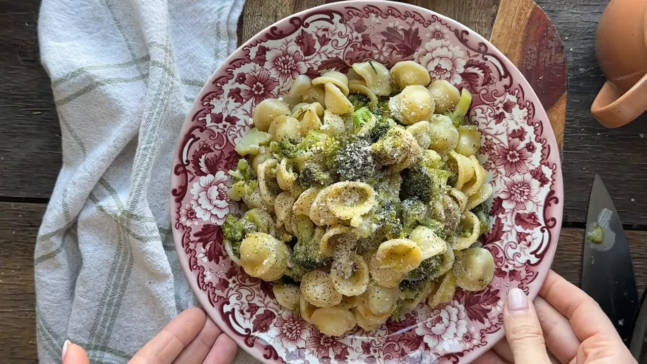 Hands presenting a bowl of orecchiette with broccoli on a floral plate, ready to serve