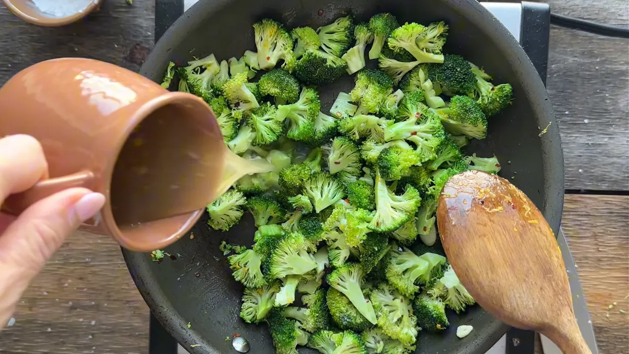 Hand pouring liquid into a skillet of chopped broccoli with a wooden spoon