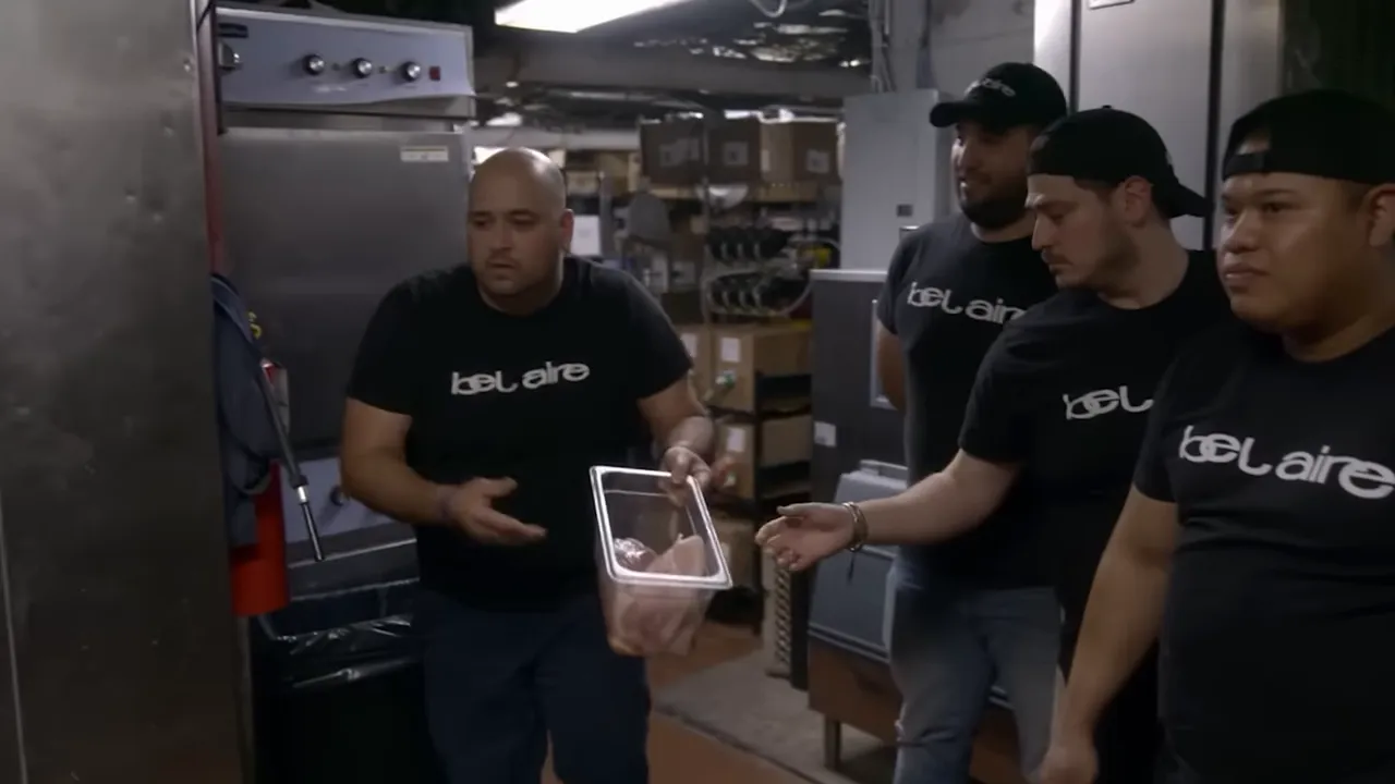 Worker holding an unsealed container of raw chicken in a cluttered walk-in refrigerator with other staff watching