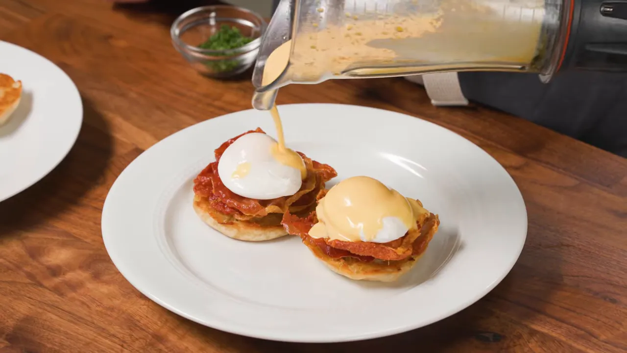 Blender hollandaise being poured over two poached eggs on English muffins on a white plate.