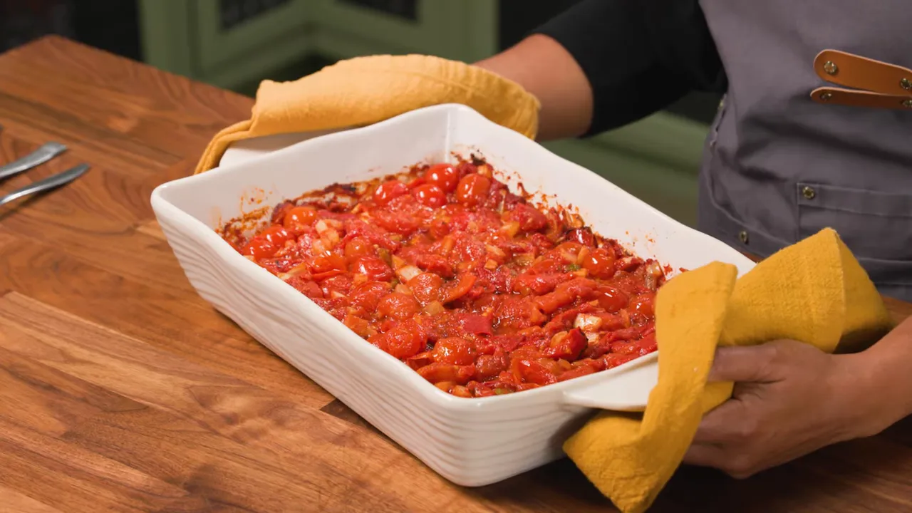 White baking dish of roasted cherry tomatoes and peppers in a red tomato sauce held with yellow oven mitts on a wooden counter