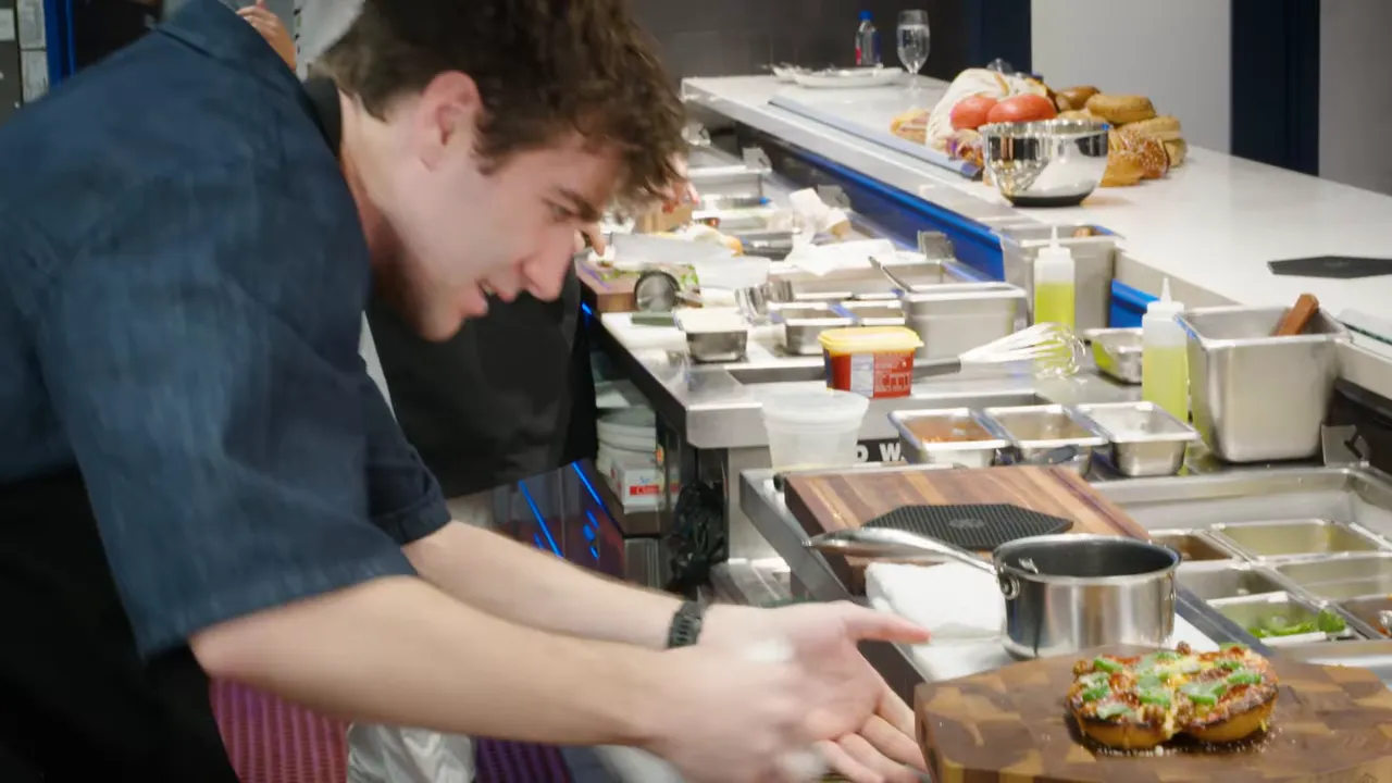 Chef plating a finished meatball-style sandwich with melted cheese on a wooden board at the pass.