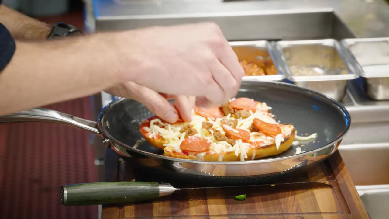 Close-up of a sub roll in a frying pan topped with marinara, shredded cheese and cured meat as hands finish the toppings.