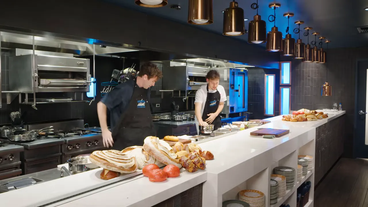 Open kitchen pass with breads and two cooks preparing ingredients on a long white counter under pendant lights.