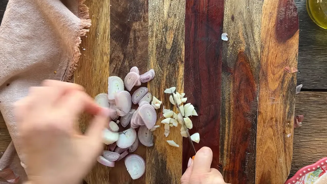 Sliced shallots and chopped garlic on a wooden cutting board with hands and knife