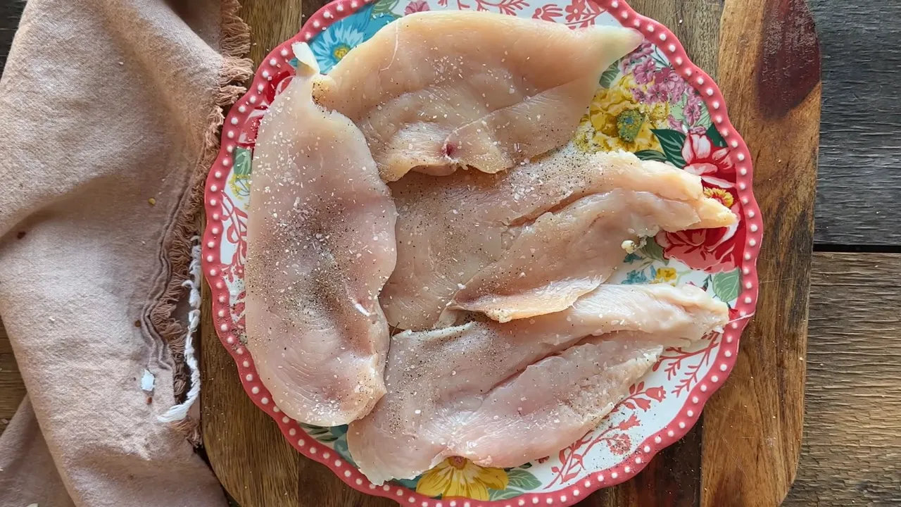 Four chicken breasts on a decorative plate seasoned with salt and pepper, ready to cook
